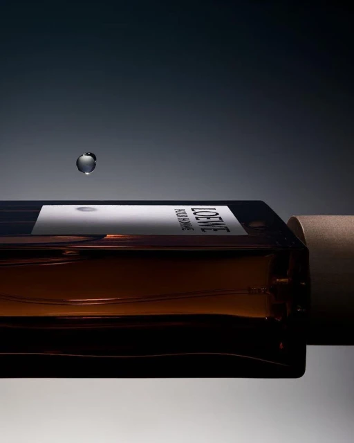 Loewe perfume bottle on its side with a water droplet hovering above it against a gradient grey and dark grey background.