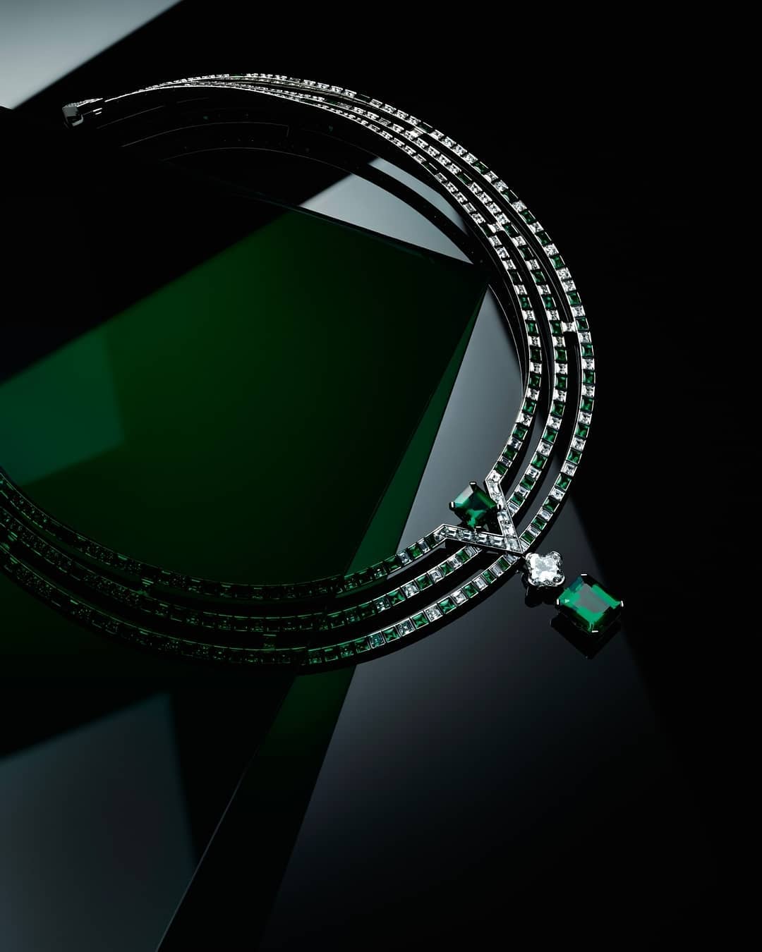 Elegant silver necklace with emerald and diamond gemstones, set against a glossy dark green and black surface for a luxurious aesthetic.
