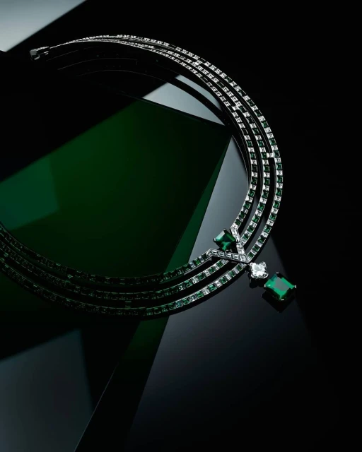Elegant silver necklace with emerald and diamond gemstones, set against a glossy dark green and black surface for a luxurious aesthetic.