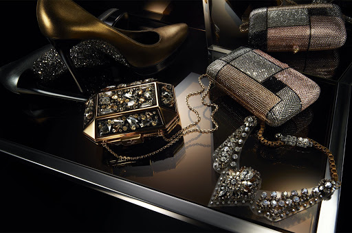 Luxury evening accessories including a gold stiletto, crystal clutches, and a sparkling diamond necklace on a reflective black surface