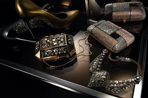 Luxury evening accessories including a gold stiletto, crystal clutches, and a sparkling diamond necklace on a reflective black surface