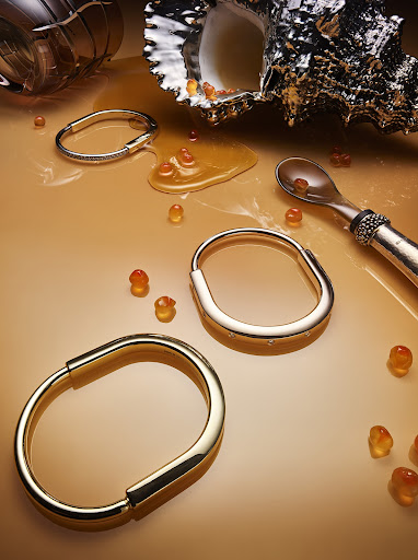 Still life studio shot of gold bracelets with spilled orange caviar beads, metallic sea shell, and spoon on a neutral background.