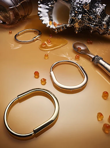 Still life studio shot of gold bracelets with spilled orange caviar beads, metallic sea shell, and spoon on a neutral background.