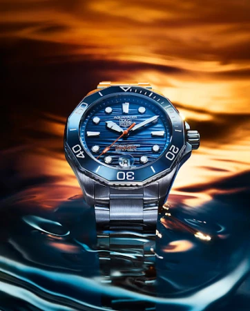 Wristwatch with a blue face and stainless-steel band on a reflective wet surface, set against a blurred orange background.