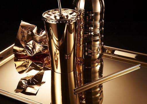 Still life of a golden plastic water bottle and a golden plastic cup with a straw, on a gold tray with black background.