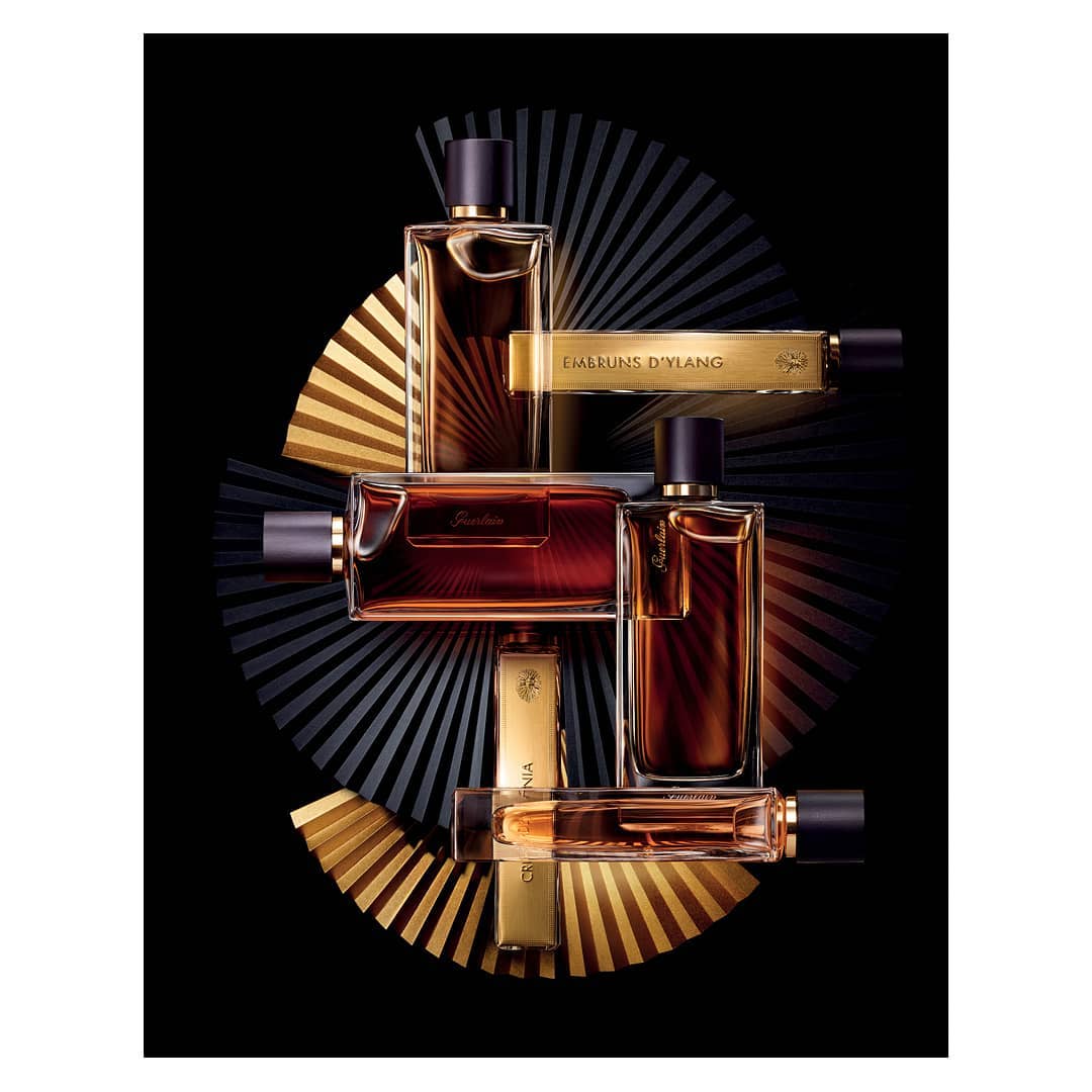 Still life of luxury Guerlain perfume bottles in amber and gold tones against a black background with a decorative radiating fan.