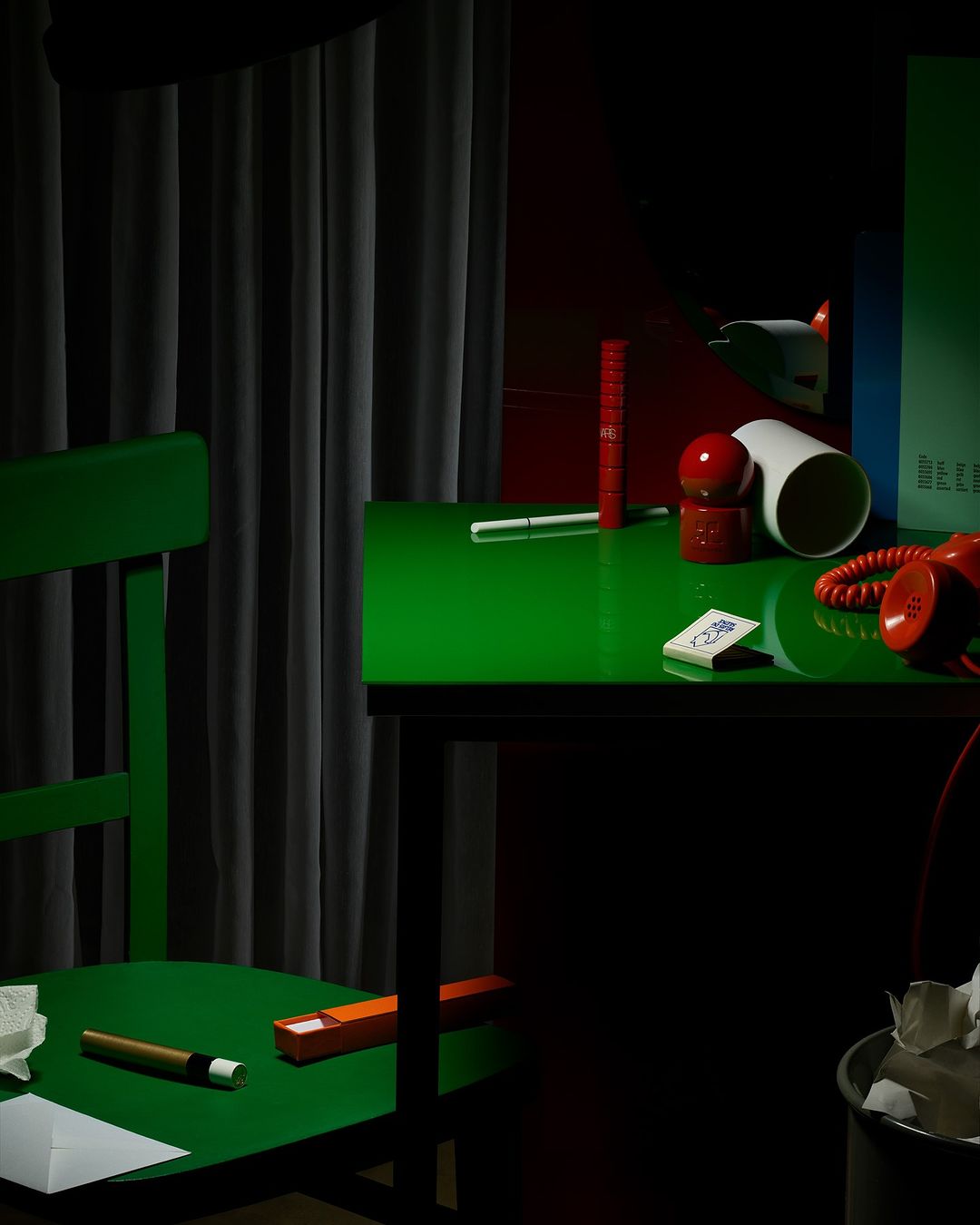Still life photograph of a green desk and chair in a room with various objects, including a red telephone and a wastepaper basket.