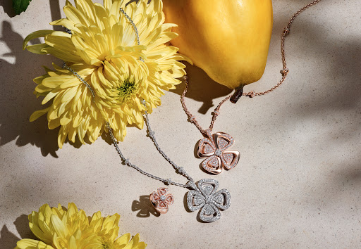 Two flower pendant necklaces alongside a yellow pear and yellow flowers on a neutral surface, creating an elegant jewelry still life.