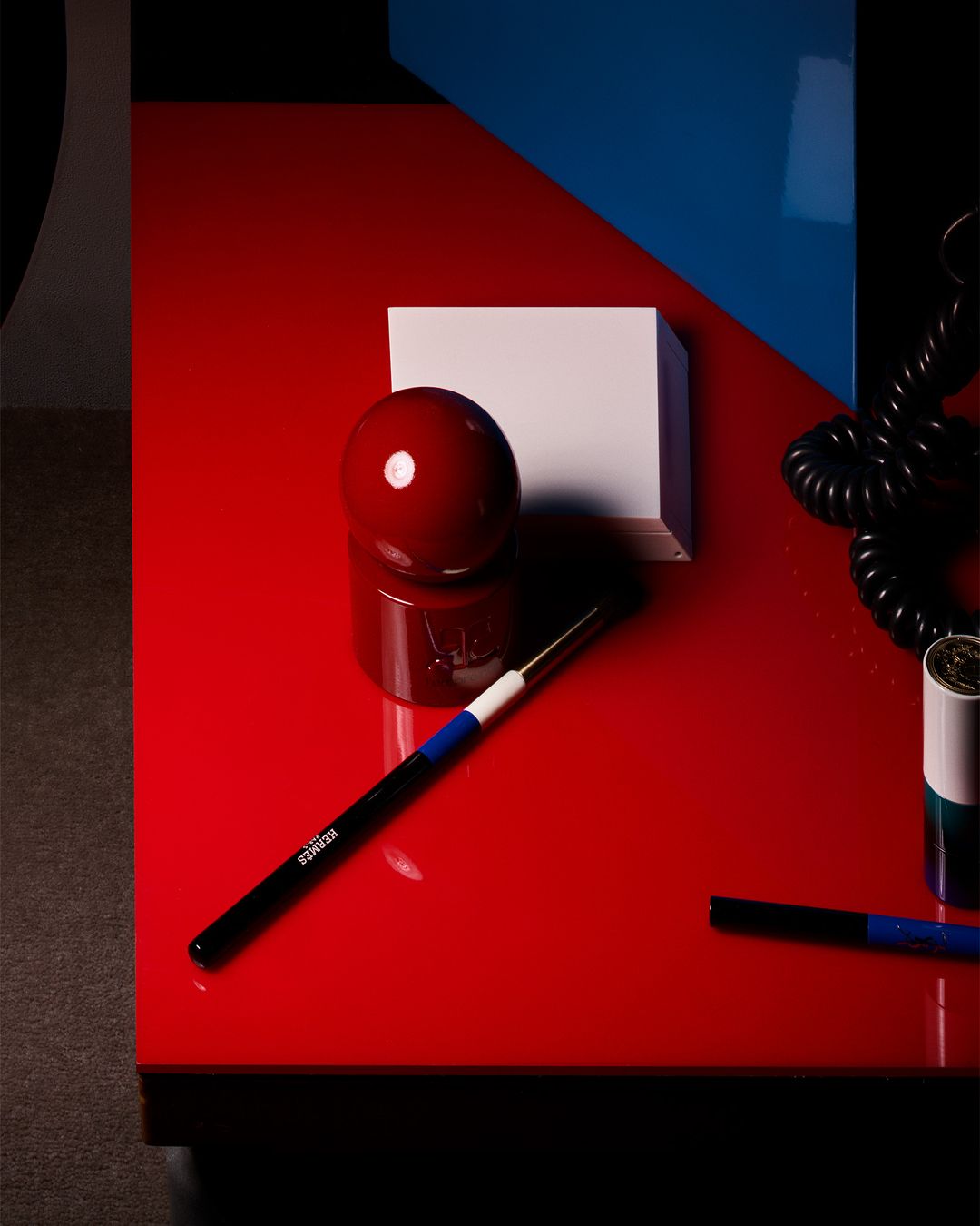 Cosmetics still life on a glossy red table with a makeup brush, lipstick, a white box, and geometric shapes in the background.