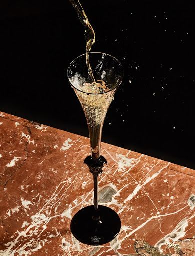 Champagne is poured into a tall stemmed glass with a black base on a marbled surface against a black background creating a moody setting.