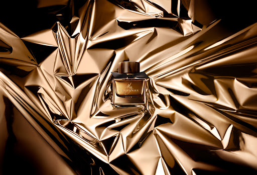 A rectangular glass perfume bottle with a dark wood cap resting on crumpled metallic gold foil background.