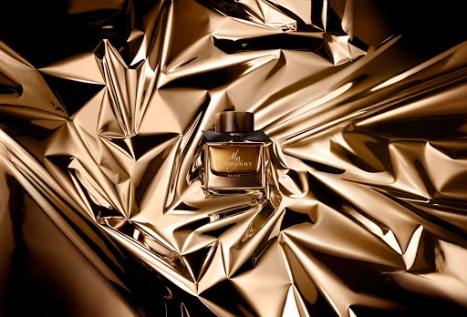 A rectangular glass perfume bottle with a dark wood cap resting on crumpled metallic gold foil background.