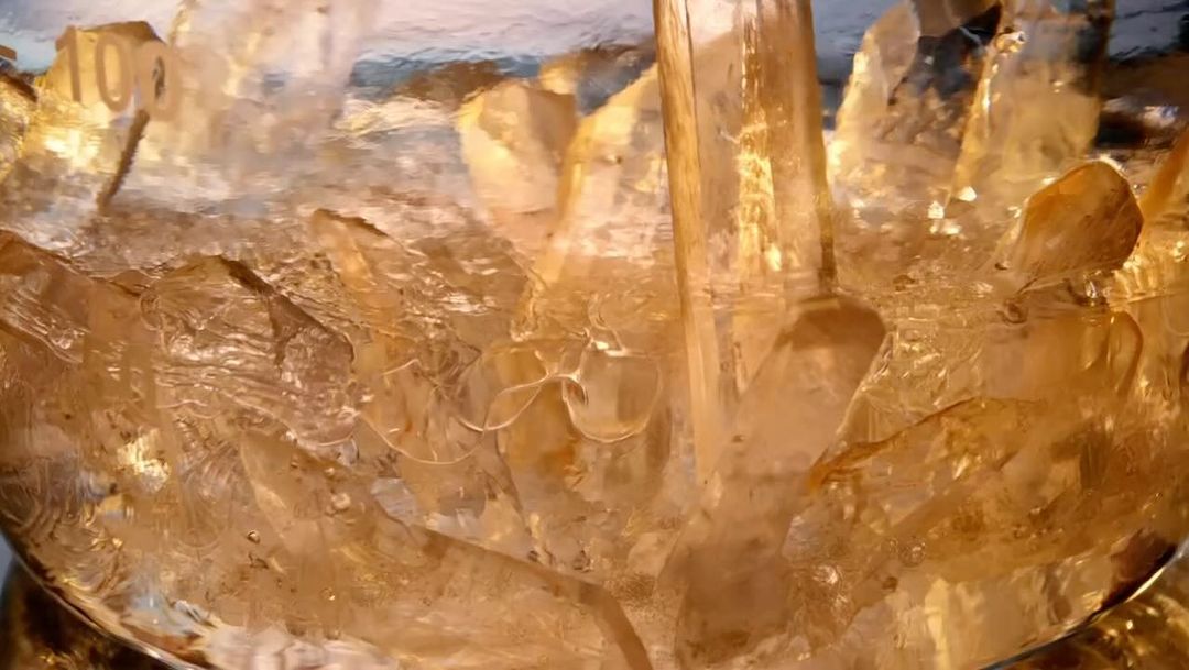Close-up shot of golden liquid being poured over ice, filling the frame with a golden hue and textural details of frozen ice.