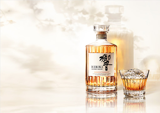 Hibiki Japanese Harmony whisky bottle and crystal glass with round ice sphere on reflective surface against light background