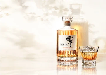 Hibiki Japanese Harmony whisky bottle and crystal glass with round ice sphere on reflective surface against light background