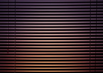 Close-up of closed horizontal window blinds with gradient light filtering through the slats in warm dark tones.