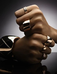 Close-up of clenched fists adorned with gold rings and a wide gold bracelet against a dark gradient background