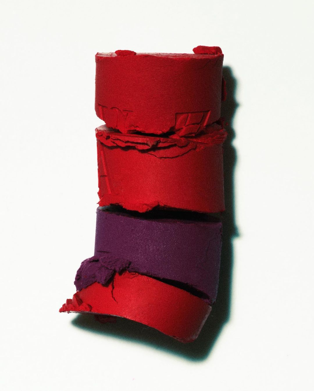 Macro photograph of a stack of cylindrical lipstick samples in red and purple hues against a clean white background with soft lighting.