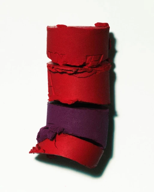 Macro photograph of a stack of cylindrical lipstick samples in red and purple hues against a clean white background with soft lighting.