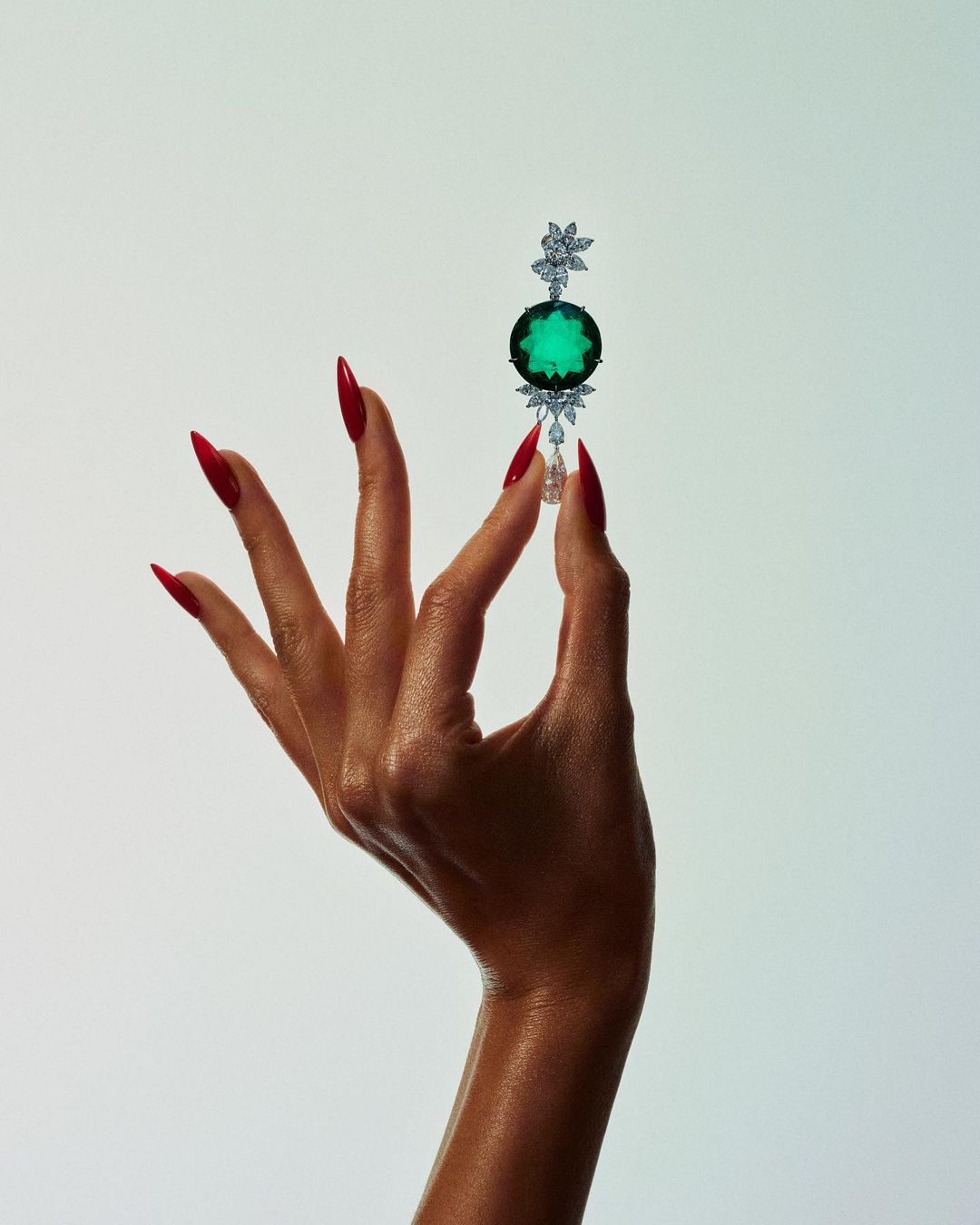 Woman's hand with red nails holds up jewelry with a large green stone on a white background in a beauty or fashion shot.