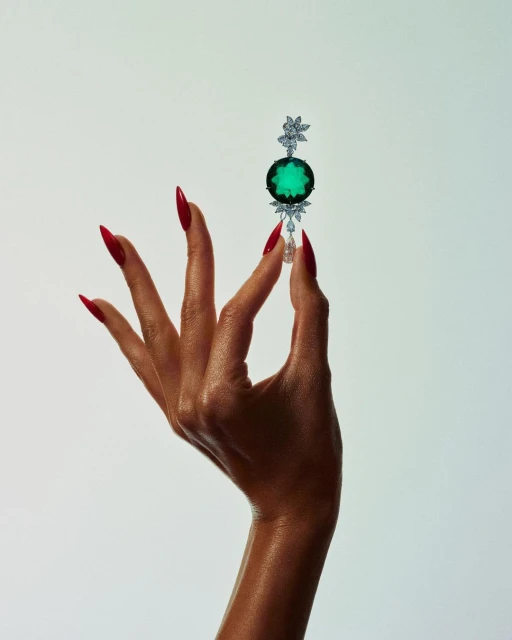 Woman's hand with red nails holds up jewelry with a large green stone on a white background in a beauty or fashion shot.