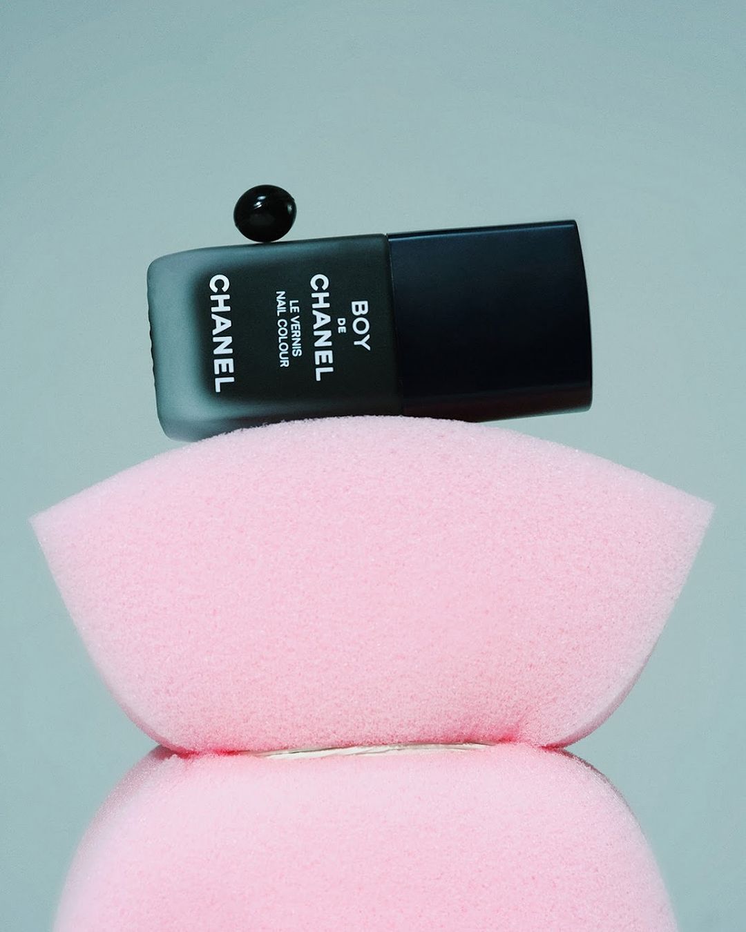 Black Chanel nail polish bottle with a floating drop on top, set on a pink makeup sponge, set against a soft grey background.