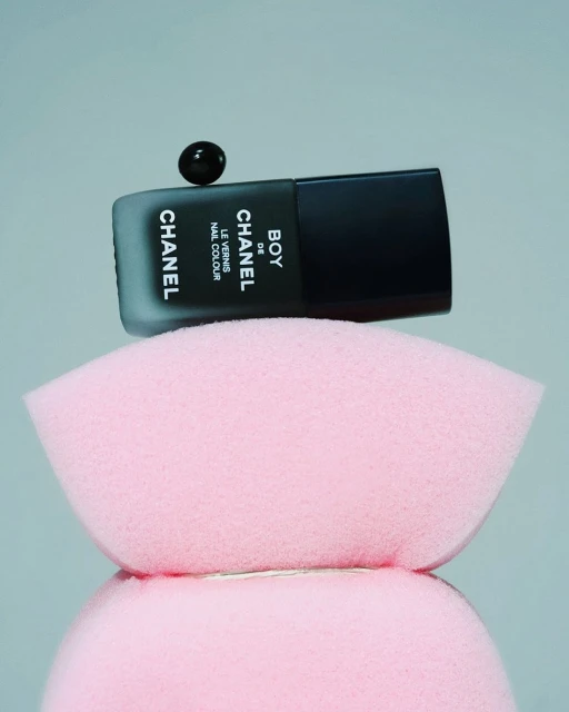Black Chanel nail polish bottle with a floating drop on top, set on a pink makeup sponge, set against a soft grey background.