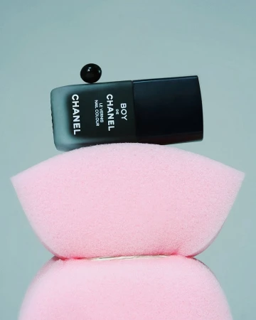 Black Chanel nail polish bottle with a floating drop on top, set on a pink makeup sponge, set against a soft grey background.