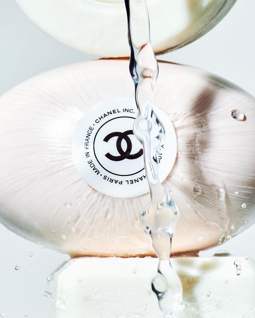 Close-up of a stack of three white Chanel soap bars with water running over them against a clean, bright background in a studio setting.