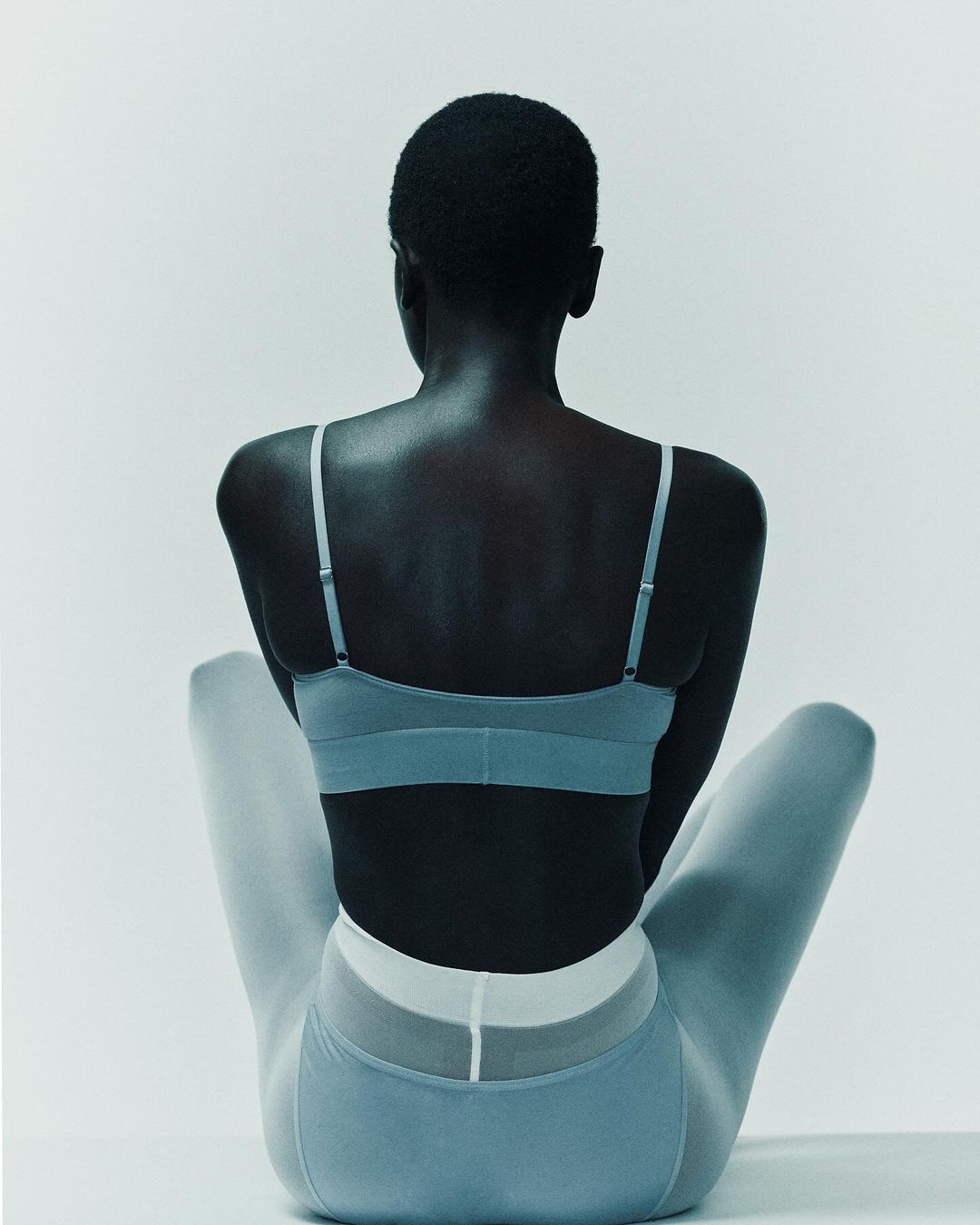 Back view of a Black woman in light blue activewear, sitting with her legs crossed against a white background, showcasing fashion and fitness.