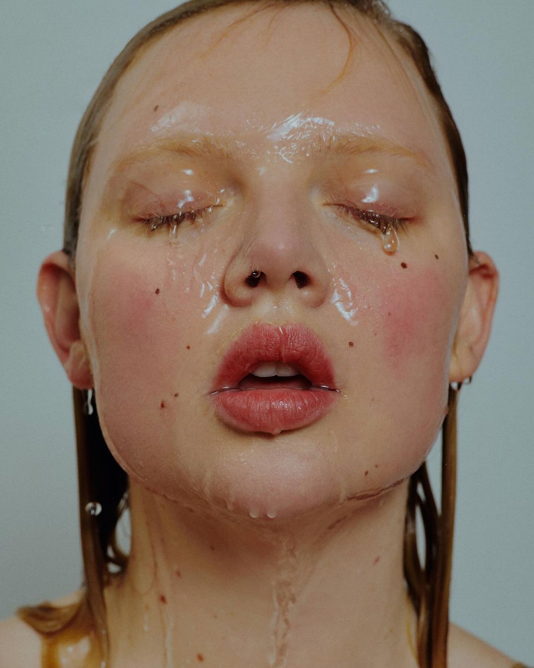Close-up of a young adult woman's face with eyes closed, water running down her face, and minimalistic makeup in a studio setting.