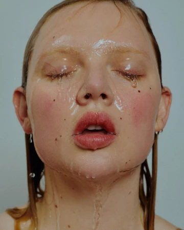 Close-up of a young adult woman's face with eyes closed, water running down her face, and minimalistic makeup in a studio setting.