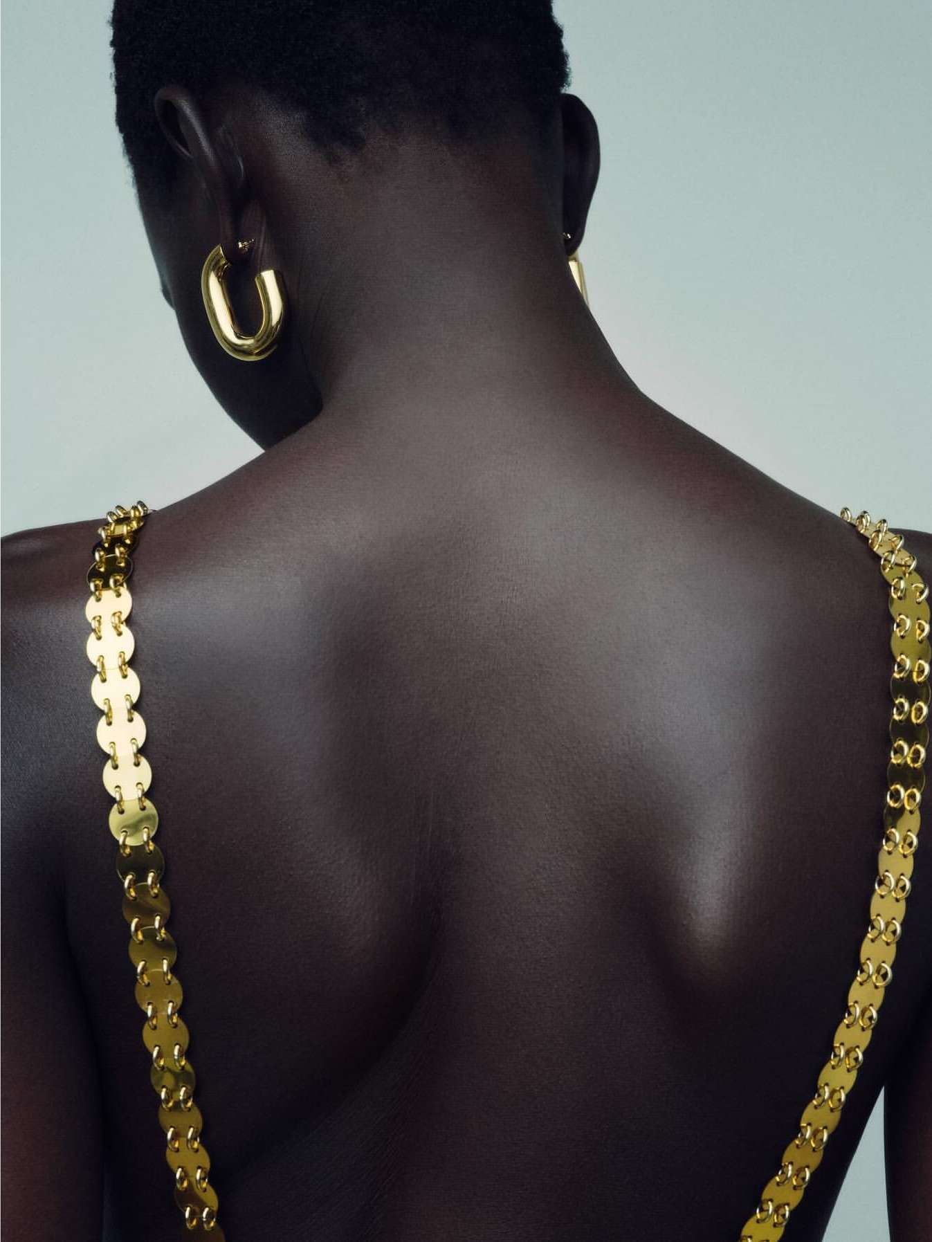 Close-up of an African-American woman's back with gold chain top and hoop earrings against a light neutral background, fashion-focused.