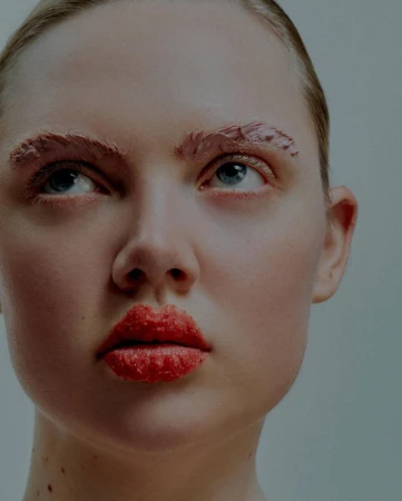 Close-up of a young fair-skinned woman with pink textured makeup on her eyebrows and lips looking up against a plain background.