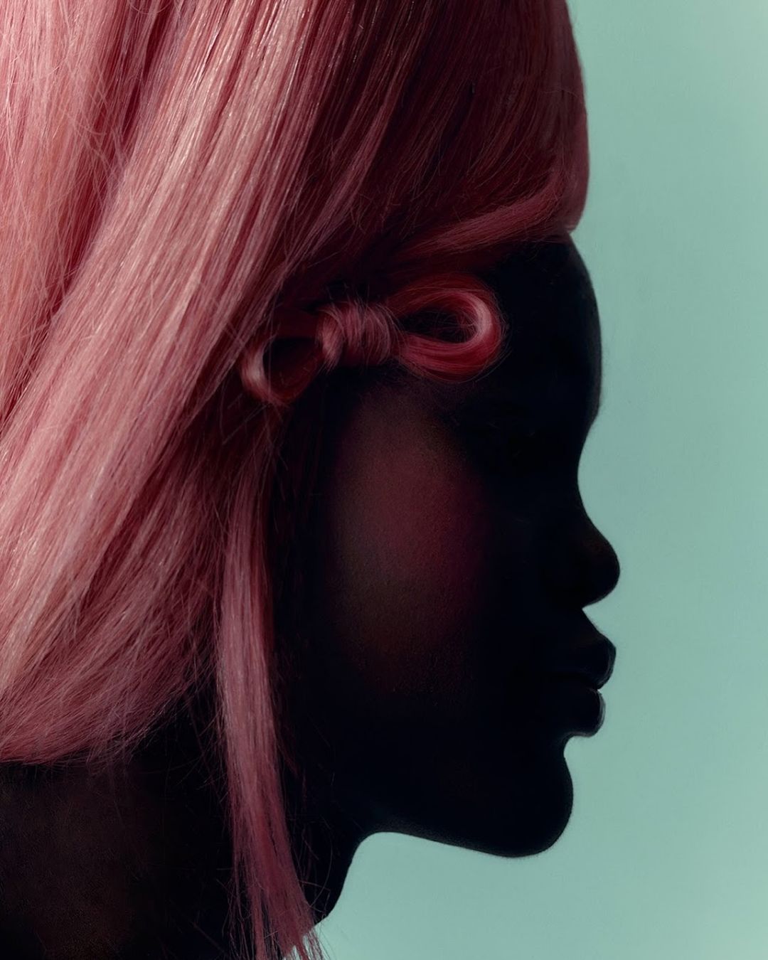 Close-up studio shot of a person's silhouette in profile with pink wig and bow detail against a light blue backdrop.