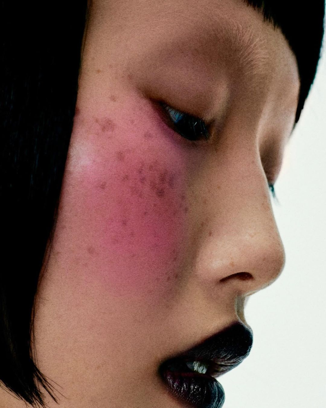 Close-up studio portrait of an Asian adult female with pink blush, black hair and dark lipstick against a white background.