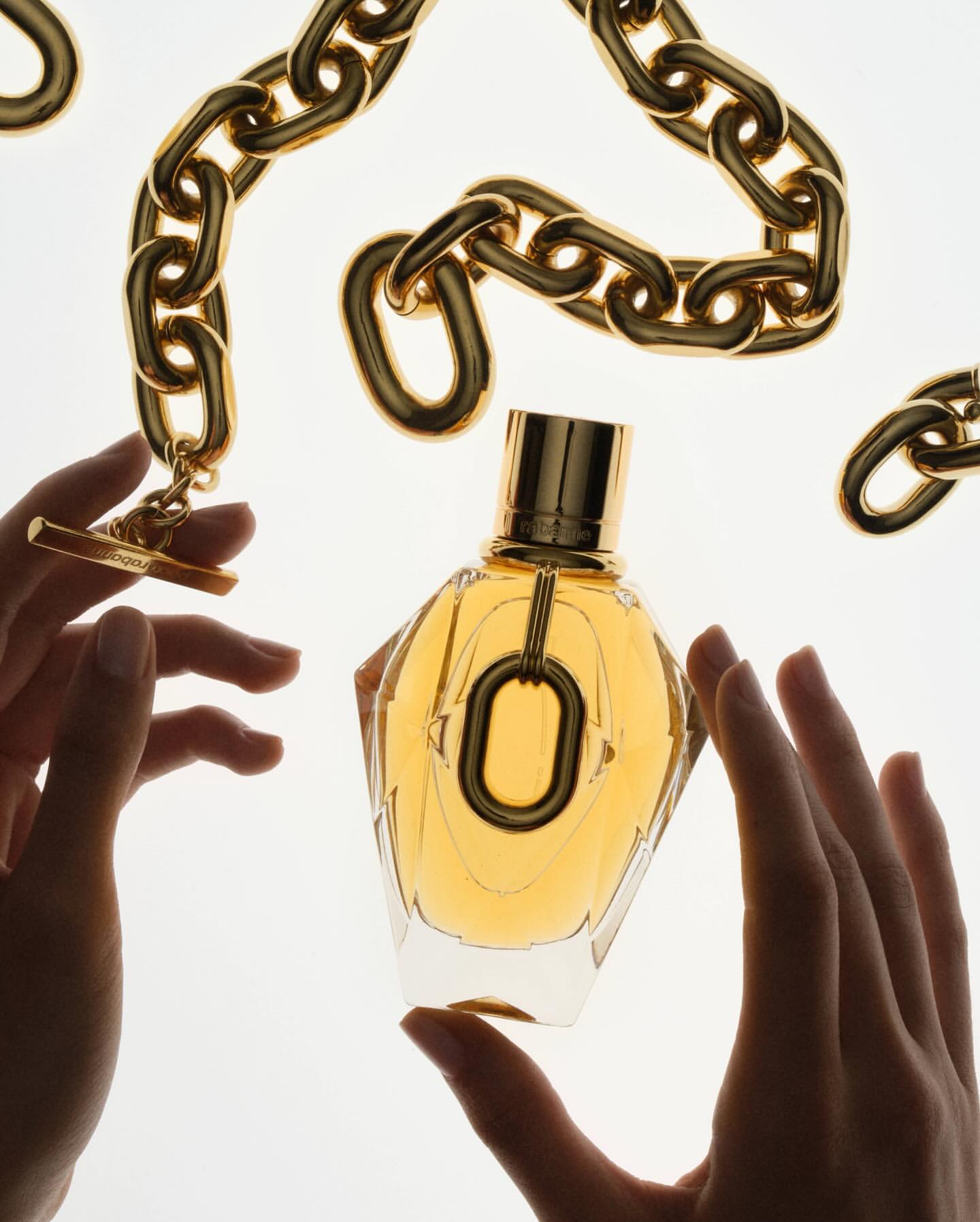 Elegant perfume bottle is held by two hands, adorned with gold chain jewelry, and set against a bright white backdrop, highlighting its design.