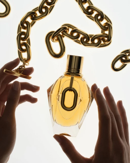 Elegant perfume bottle is held by two hands, adorned with gold chain jewelry, and set against a bright white backdrop, highlighting its design.
