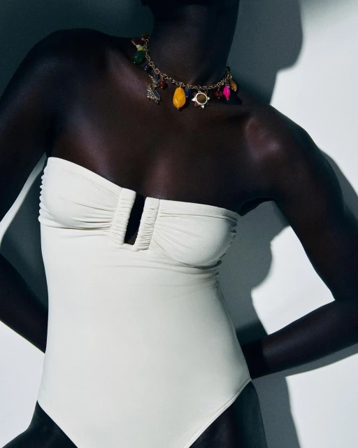 A woman wearing a cream-colored strapless swimsuit and a colorful charm necklace is standing in front of a white background in a studio.