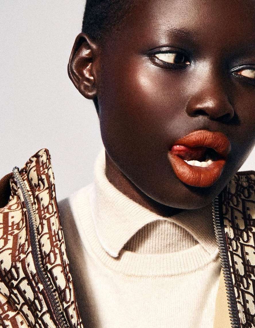 Close-up shot of a dark-skinned woman with short hair wearing a turtleneck and patterned jacket, sticking her tongue out slightly.