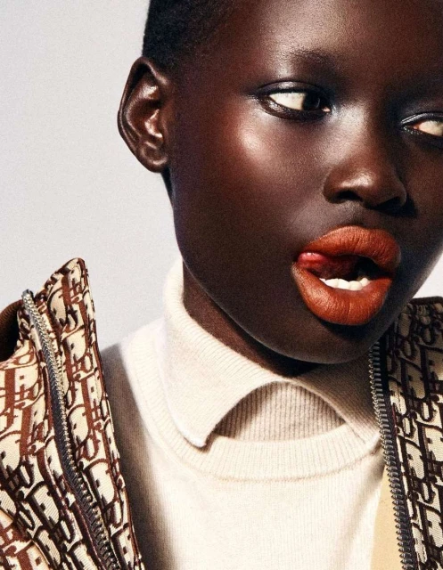 Close-up shot of a dark-skinned woman with short hair wearing a turtleneck and patterned jacket, sticking her tongue out slightly.