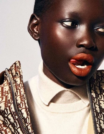Close-up shot of a dark-skinned woman with short hair wearing a turtleneck and patterned jacket, sticking her tongue out slightly.