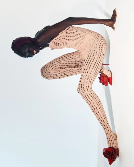 Fashion editorial shot of a dark-skinned model in a light pink patterned bodysuit, red rose heels, and head covering posing against a wall.