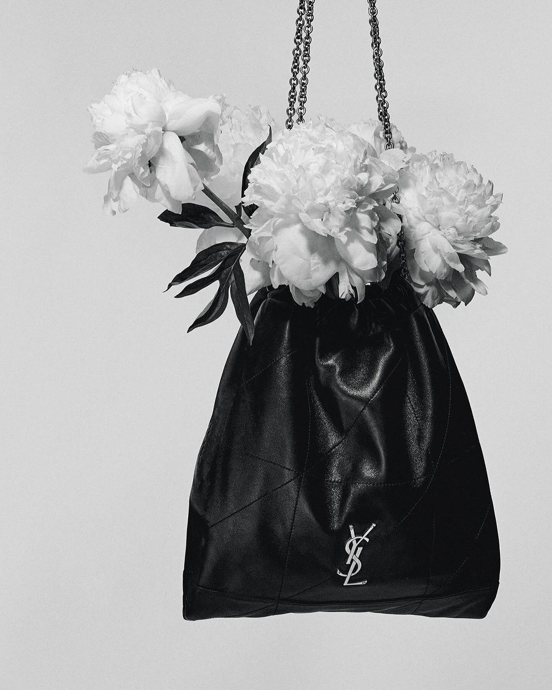 Black leather bucket bag with silver chain and signature logo filled with white peonies in a studio against a white background.