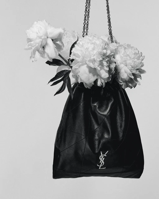 Black leather bucket bag with silver chain and signature logo filled with white peonies in a studio against a white background.