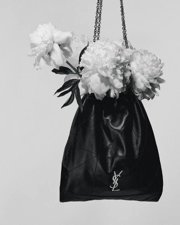 Black leather bucket bag with silver chain and signature logo filled with white peonies in a studio against a white background.