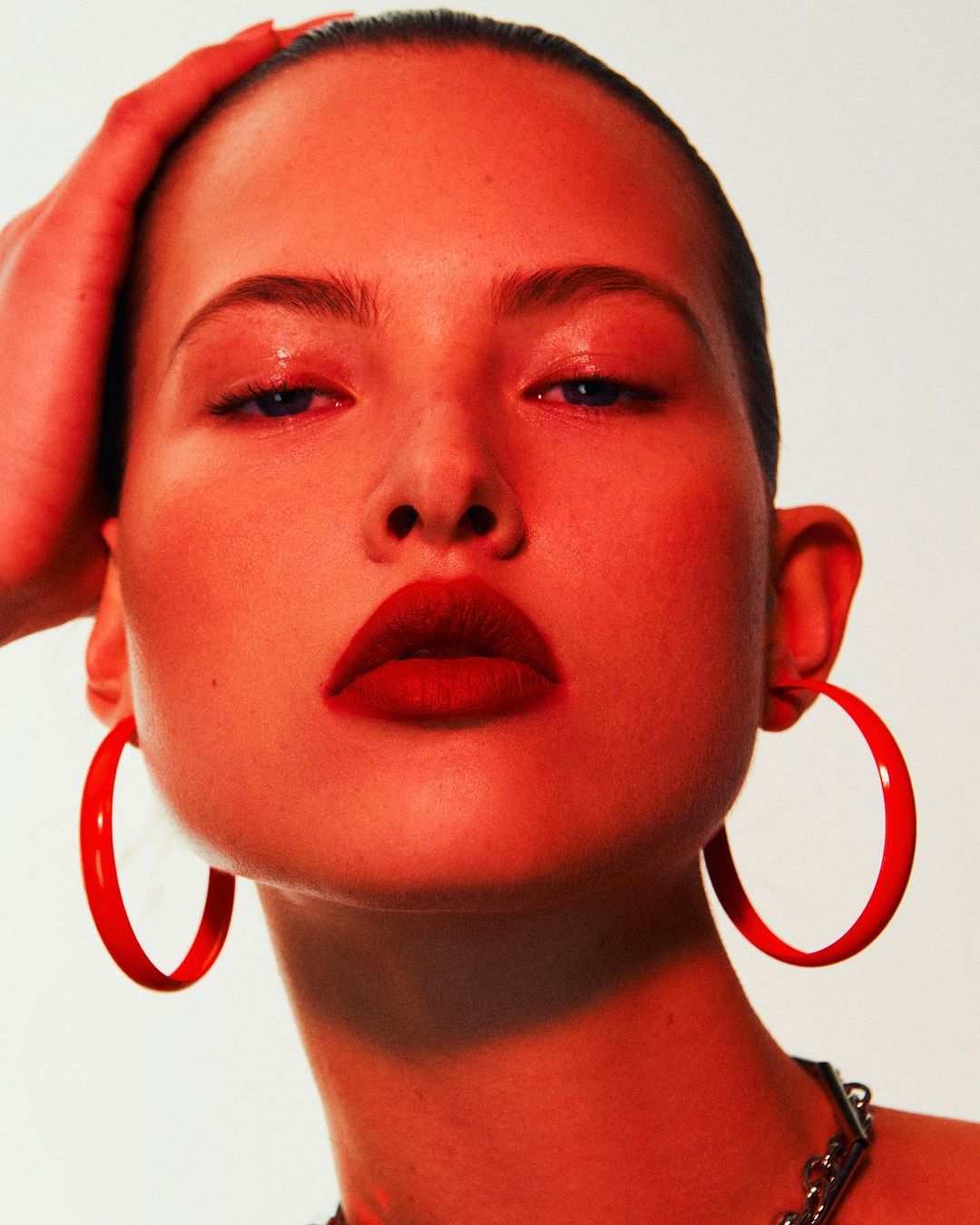 Close-up portrait of a woman with a shaved head wearing large red hoop earrings and dark lipstick under red lighting.