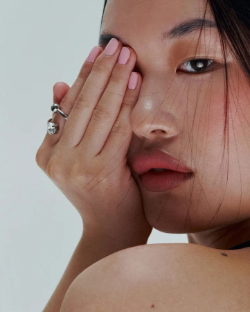 Close-up portrait of a woman holding her hand across her face with pink nail polish and silver rings against a plain background