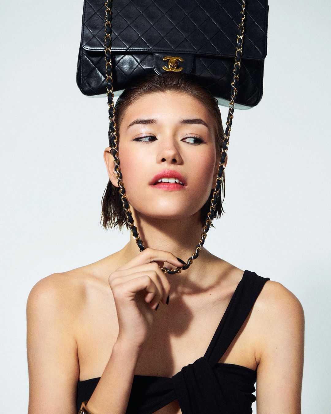 Woman poses in a studio with a black Chanel handbag balanced on her head and the chain draped around her neck against a white backdrop.