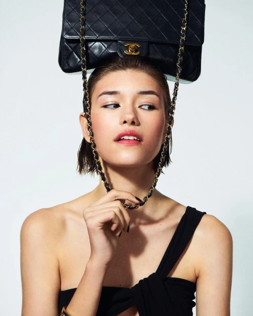 Woman poses in a studio with a black Chanel handbag balanced on her head and the chain draped around her neck against a white backdrop.